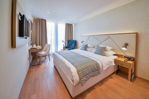 Habitación de hotel con cama, mesita de noche, lámpara, silla, escritorio y televisor.