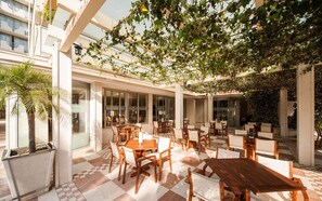 An outdoor cafe with tables and chairs, a palm tree, and a building with a restaurant sign. - Mediteran Wellness & Spa, Congress Center (Bečići)