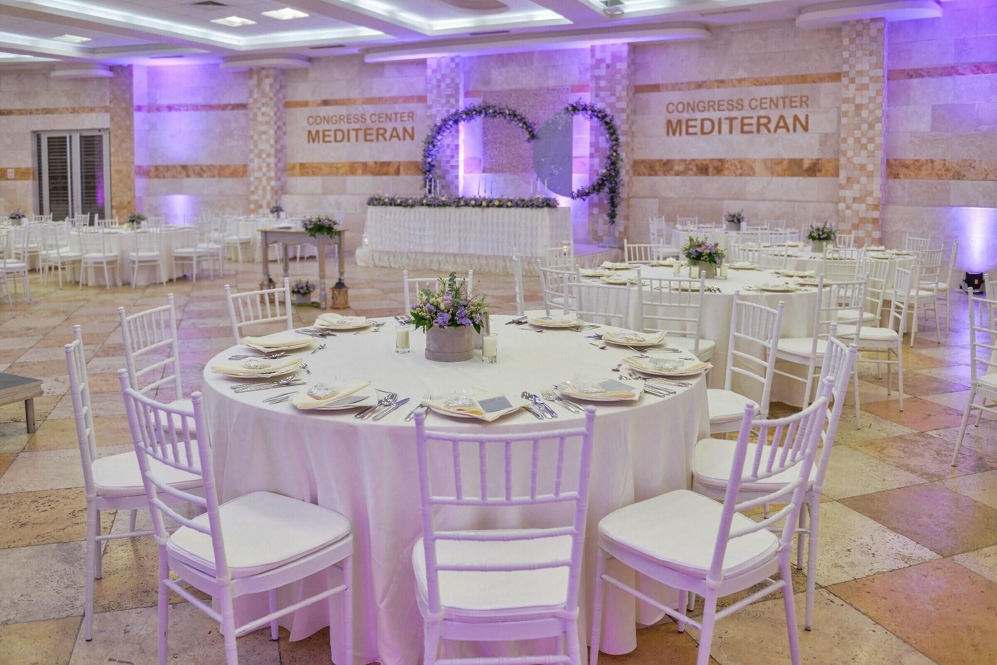 Un centro de convenciones con mesas redondas y sillas blancas, suelo de baldosas y una pared con el rótulo 'CONGRESS CENTER MEDITERAN'.