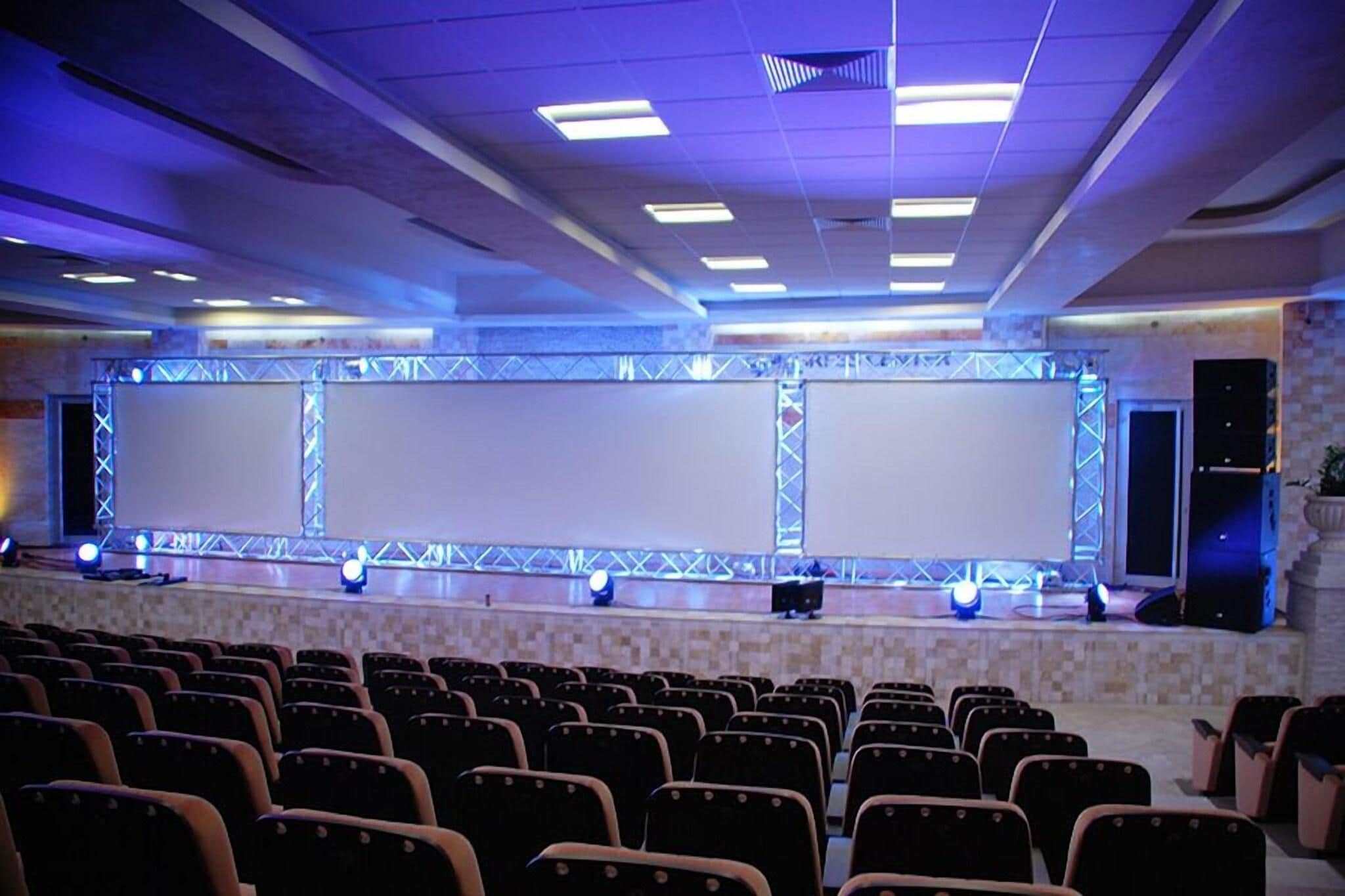 Un salón de conferencias con un escenario, varias pantallas y filas de sillas.