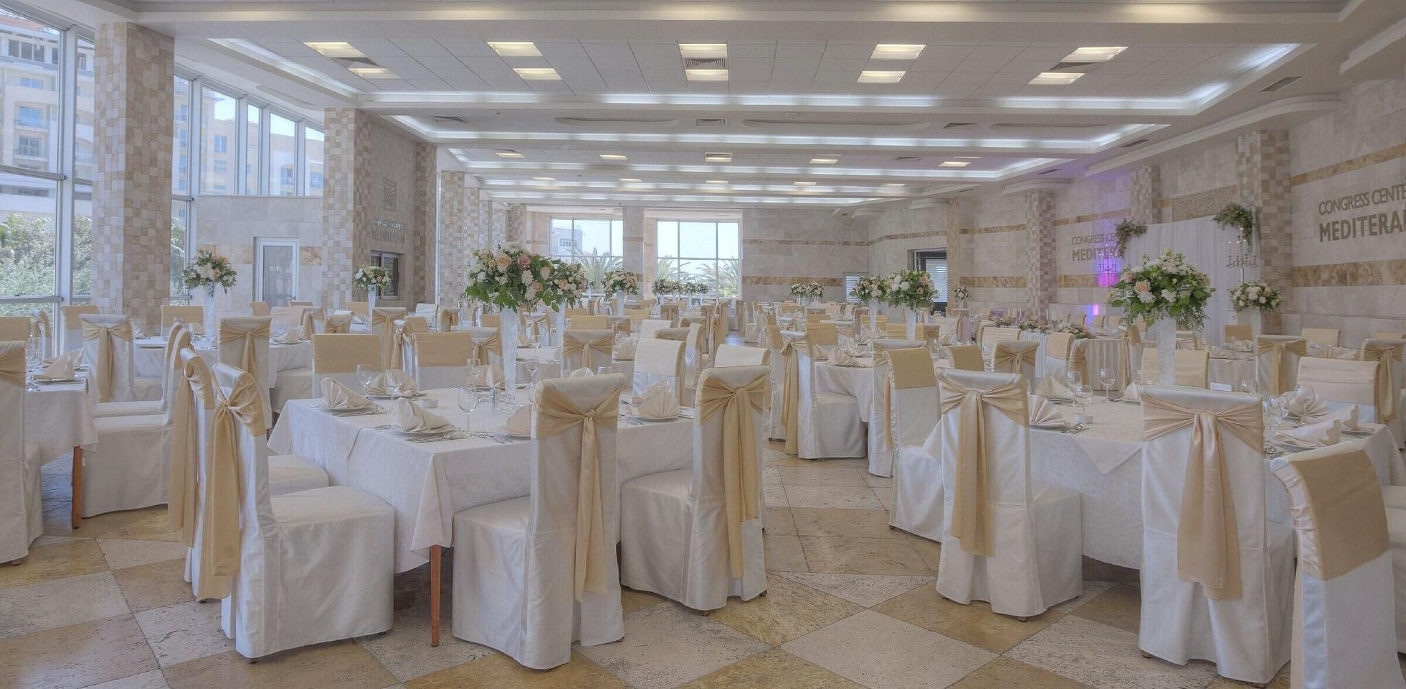 Une vaste salle de banquet, avec de longues rangées de tables, chacune entourée de chaises recouvertes d’un tissu beige et ornées de nœuds. La pièce est dotée d’un haut plafond, d’un éclairage encastré et de grandes fenêtres.