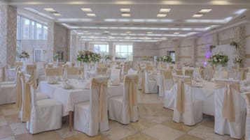 Une vaste salle de banquet, avec de longues rangées de tables, chacune entourée de chaises recouvertes d’un tissu beige et ornées de nœuds. La pièce est dotée d’un haut plafond, d’un éclairage encastré et de grandes fenêtres.