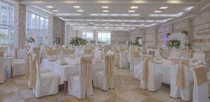 Un gran salón de banquetes con filas de mesas, cada una con sillas cubiertas de tela color beige y atadas con lazos. La sala tiene un techo alto con iluminación empotrada y ventanas amplias.