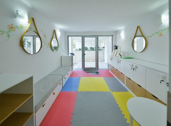 Un'area giochi per bambini con pavimento in schiuma colorato, mobili contenitori bianchi e specchi alle pareti.