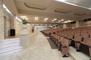 Un salón de conferencias con filas de sillas, un escenario con un atril y un pilar de piedra con una planta.