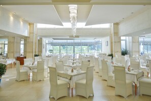 A spacious dining area with high ceilings, large windows, and a chandelier. Tables are set with white tablecloths and chairs. - Mediteran Wellness & Spa, Congress Center (Bečići)