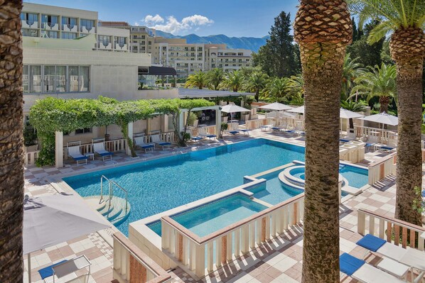 A swimming pool with lounge chairs and umbrellas, surrounded by palm trees and buildings. - Mediteran Wellness & Spa, Congress Center (Bečići)