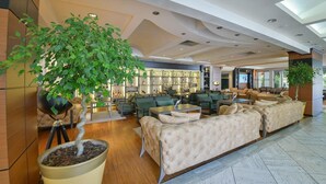 A spacious lobby with a large potted tree, seating areas, and a bar with shelves of bottles. - Mediteran Wellness & Spa, Congress Center (Bečići)
