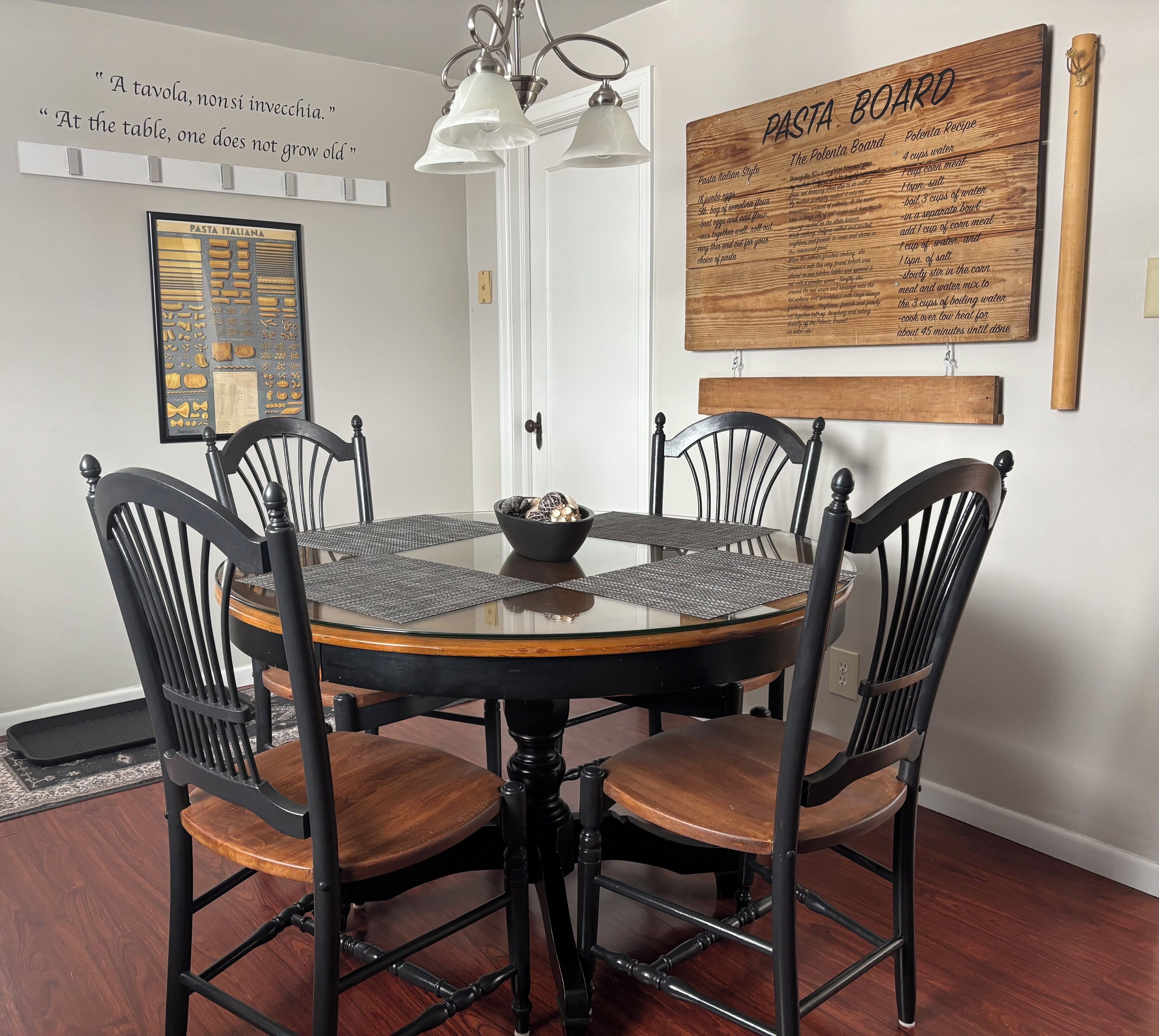 A round dining table with black chairs, a framed pasta recipe board, and a wall-mounted light fixture.