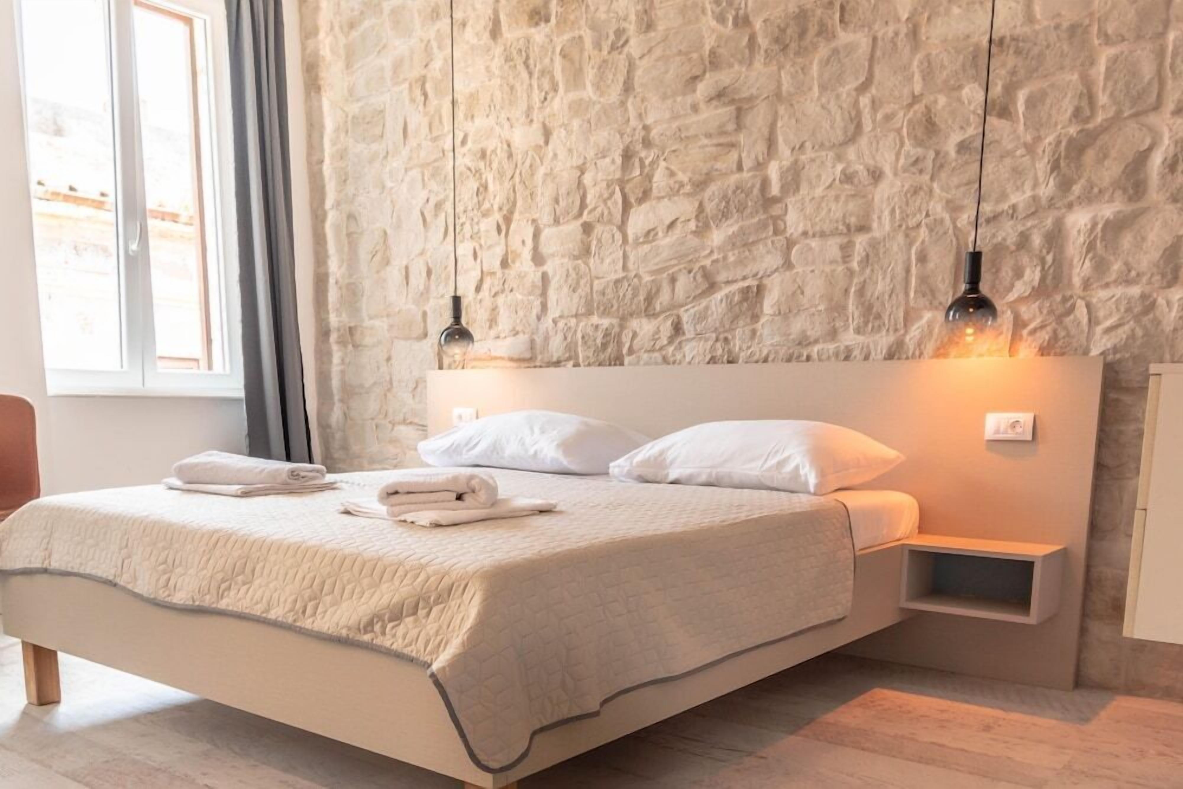 A bedroom with a stone wall, a bed with white bedding, two pendant lights, and a nightstand.