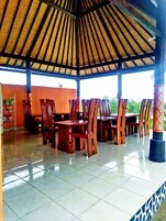Un espacio cubierto para comer al aire libre con mesas y sillas de madera, piso de baldosas y techo de paja.