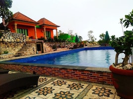 Una piscina con un borde de baldosas, un muro de piedra y una casa con tejado rojo.