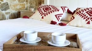 Una cama con una bandeja de madera que sostiene dos tazas y platillos blancos, frente a una pared de piedra y un cabecero blanco.