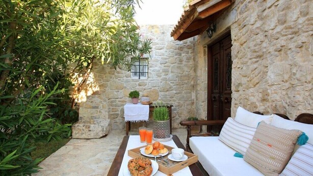 Un área de estar al aire libre con paredes de piedra y una mesa de madera dispuesta para el desayuno, con pan, pasteles y bebidas.