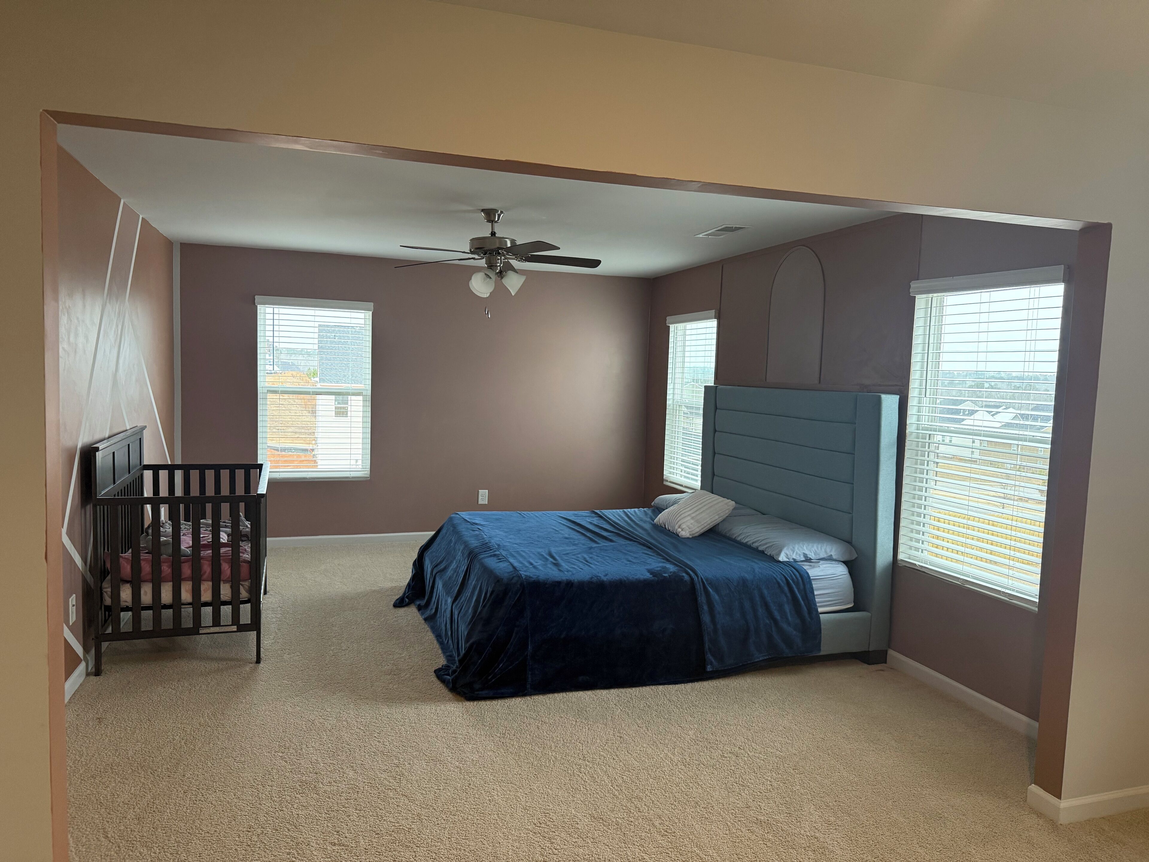 A bedroom with a bed, a crib, a ceiling fan, and large windows.