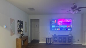 Sala de estar con televisor de pantalla plana montado en la pared, mueble de madera para entretenimiento, ventilador de techo y un pasillo que conduce a una puerta.
