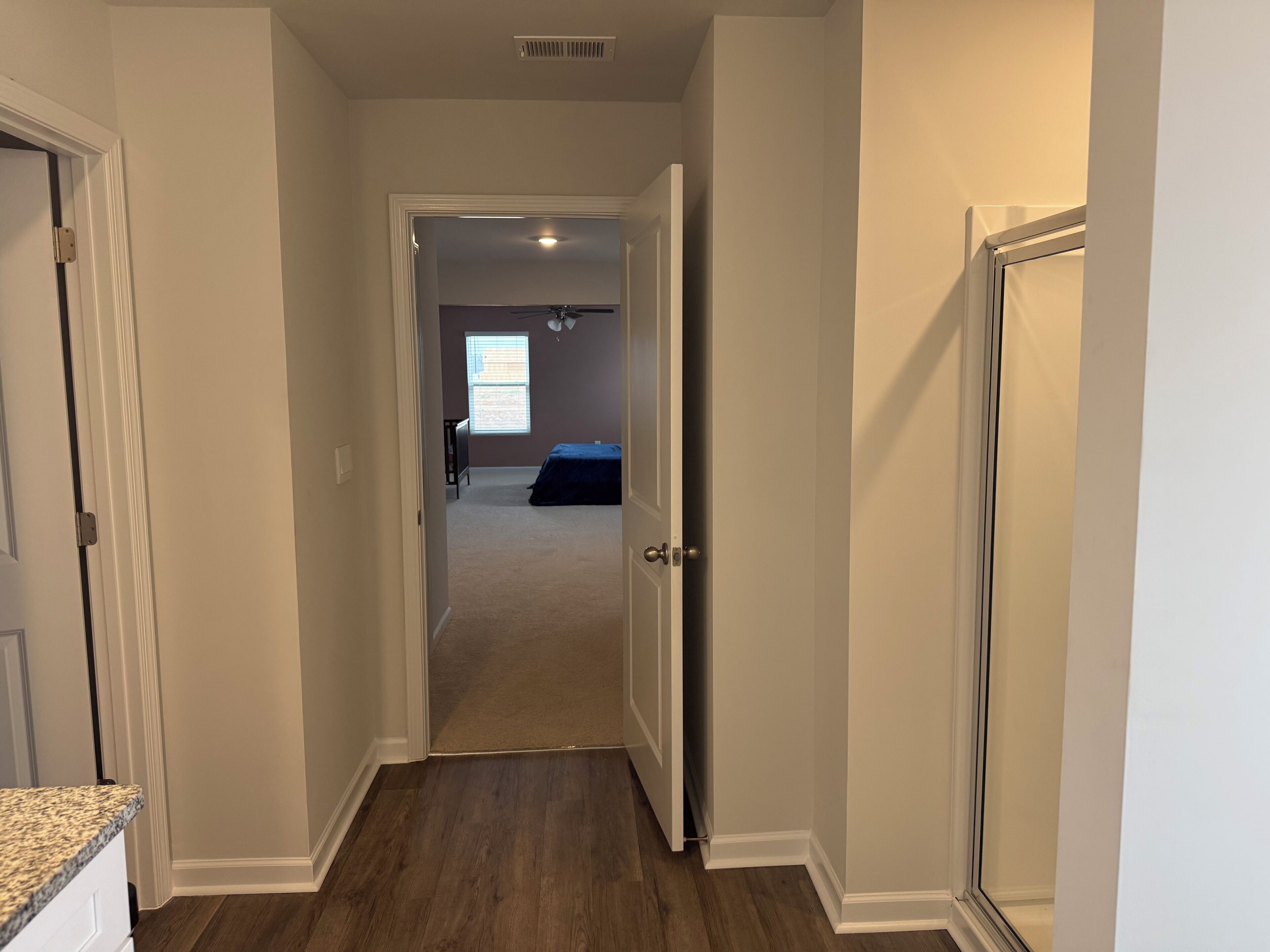 A hallway with carpeted flooring, a ceiling vent, and a door leading to a bedroom with a bed and a window.