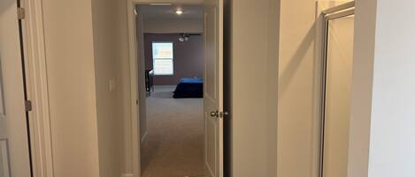 A hallway with carpeted flooring, a ceiling vent, and a door leading to a bedroom with a bed and a window.