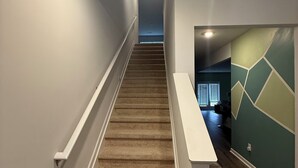 Una escalera alfombrada con barandillas blancas que conduce a una habitación con pared de diseño geométrico y una ventana con persianas.
