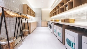 A modern laundry room with a central washing machine and dryer, wooden shelves with baskets, and a clothes drying rack with baskets. - Atour Hotel (Hangzhou)