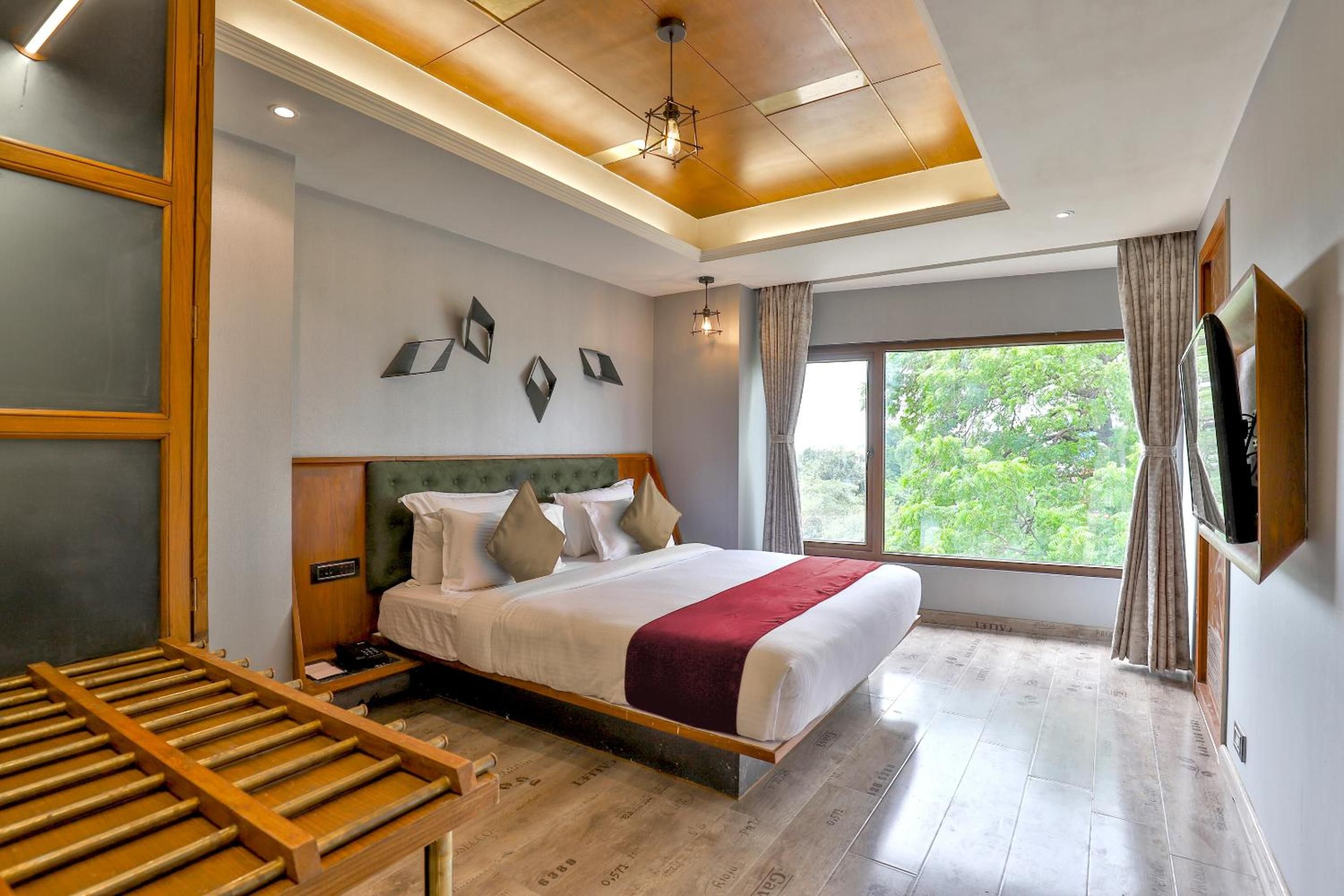 A hotel room with a bed, a wooden desk, a television, and a window with a view of trees.