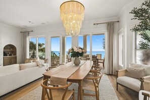 Ein geräumiges Wohnzimmer mit einem Holzespeisetisch, weißen Sofas und einem großen Fenster mit Meerblick.