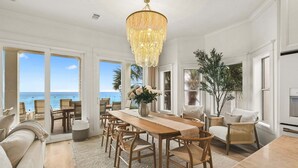 A spacious dining area with a wooden table and chairs, a chandelier, and large windows offering a view of the ocean. - GULF-FRONT w/ Private Beach! Private Pool, Hot Tub! Sleeps 24! (Rosemary Beach)