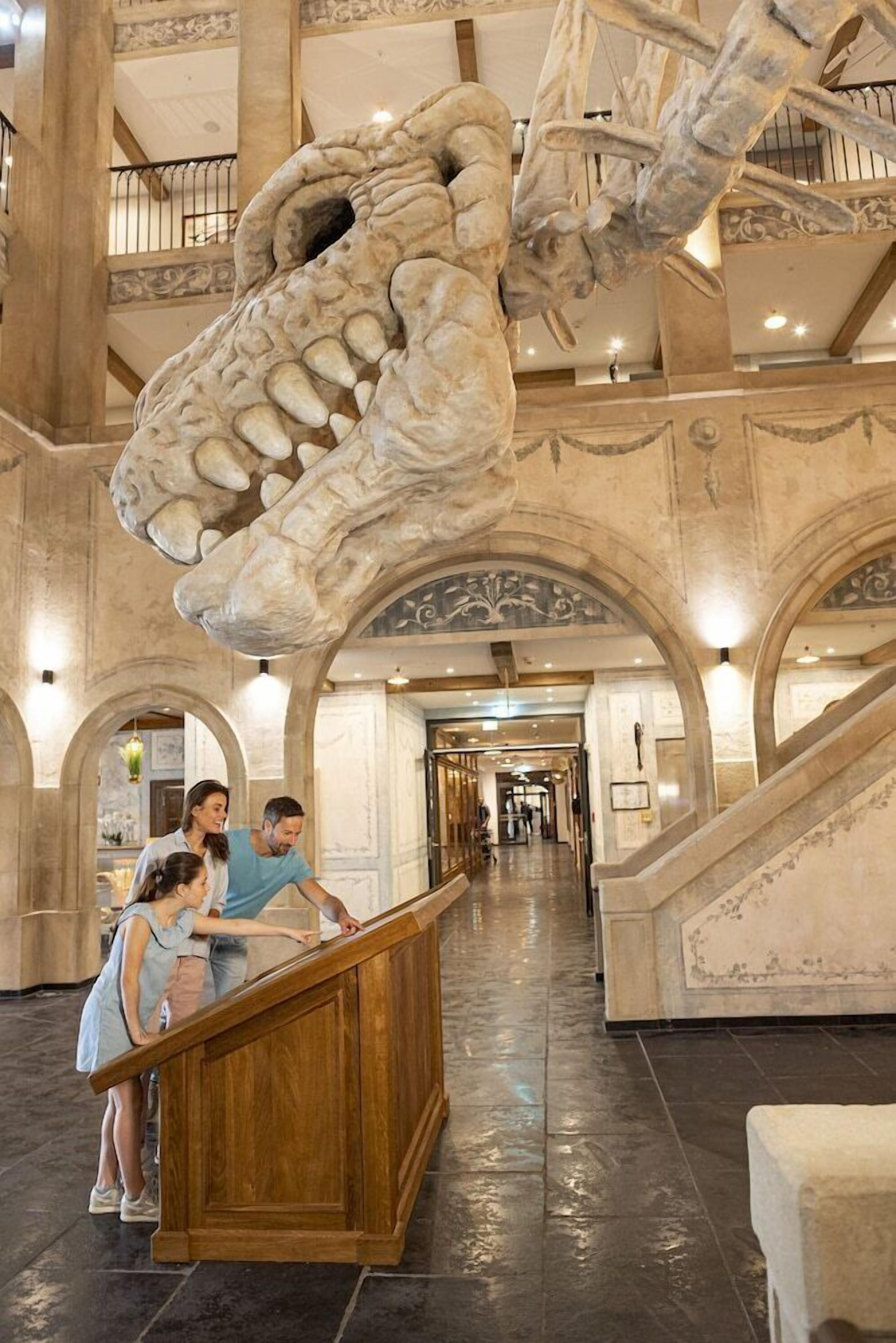 Un vaste hall d’entrée orné d’une imposante sculpture représentant le squelette d’un dinosaure, d’un escalier en bois et d’un plafond élevé agrémenté de moulures complexes.