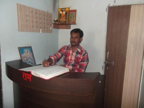 A man sitting at a wooden desk with a book and pen, a framed picture, and a shelf with a statue. - Hotel Oasis (Daund)