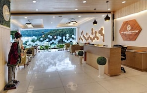 A hotel lobby with a reception desk, seating area, and a large window with a view of trees. - The Orchard Greens Resort (Manali)
