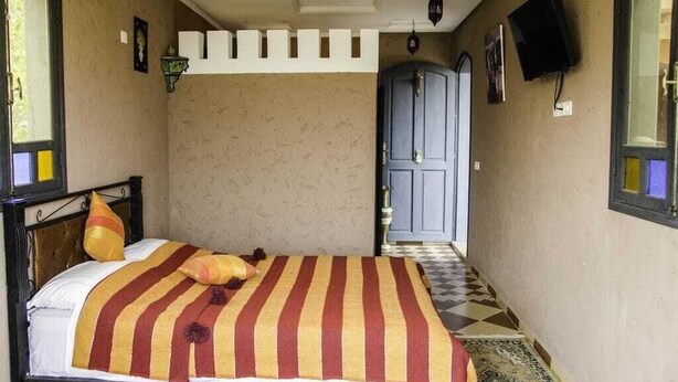 A narrow hallway with a bed on the left, a blue door at the end, and a mounted TV on the wall. - Dar Soulaimane Ourika (Marrakech)