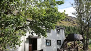 A white building with a wooden staircase leading to a platform, surrounded by trees and a hillside in the background. - Reynivellir (Reynivellir)