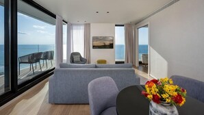 A modern living room with a large grey sofa, a small round table with a vase of flowers, and a view of the ocean through large windows. - West Lagoone Hotel (Netanya)