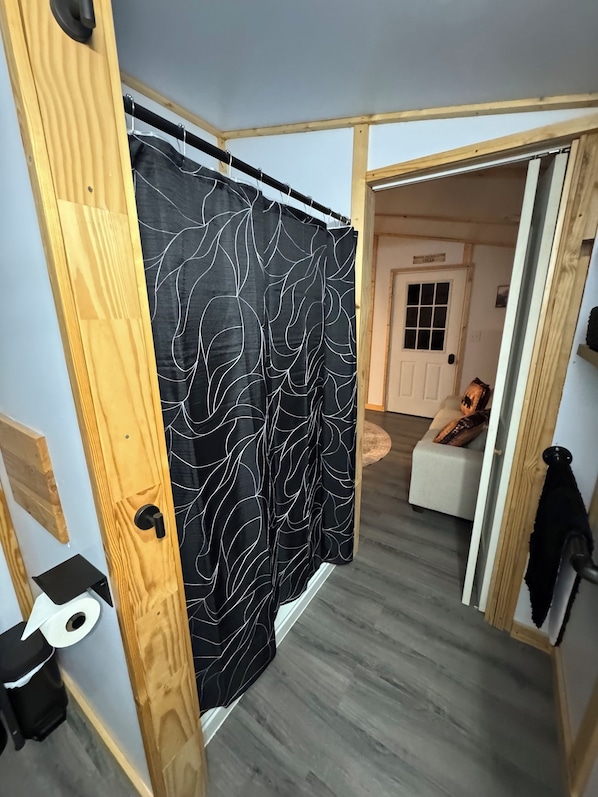 Une salle de bain avec une porte en bois, un rideau noir orné de motifs de feuilles blanches, des toilettes blanches et un sol gris.