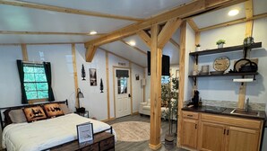 A bedroom with a bed, a nightstand, a dresser, a wooden ceiling, and a kitchen area with a sink and shelves. - cozy one-bed, one-bath cabin in Galeton, PA. Pine Creek within walking distance (Galeton)