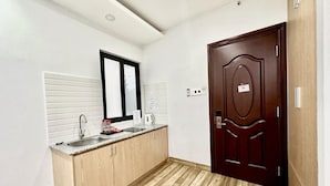 Una cocina pequeña y moderna con encimera de madera, un fregadero y un espejo encima. A la derecha se ve una puerta de madera oscura y una pared de color claro con una ventana.