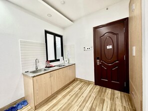 Una cocina pequeña y moderna con encimera de madera, un fregadero y un espejo encima. A la derecha se ve una puerta de madera oscura y una pared de color claro con una ventana.
