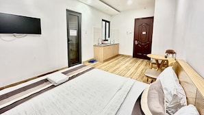 Habitación de hotel con cama, televisión montada en la pared, una pequeña cocina americana, piso de madera y una puerta que da a otra habitación.