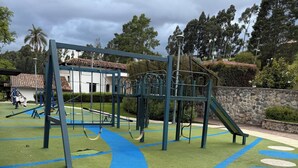 Un terrain de jeux doté d’une structure d’escalade et d’un toboggan, entouré d’un mur de pierre et de verdure.