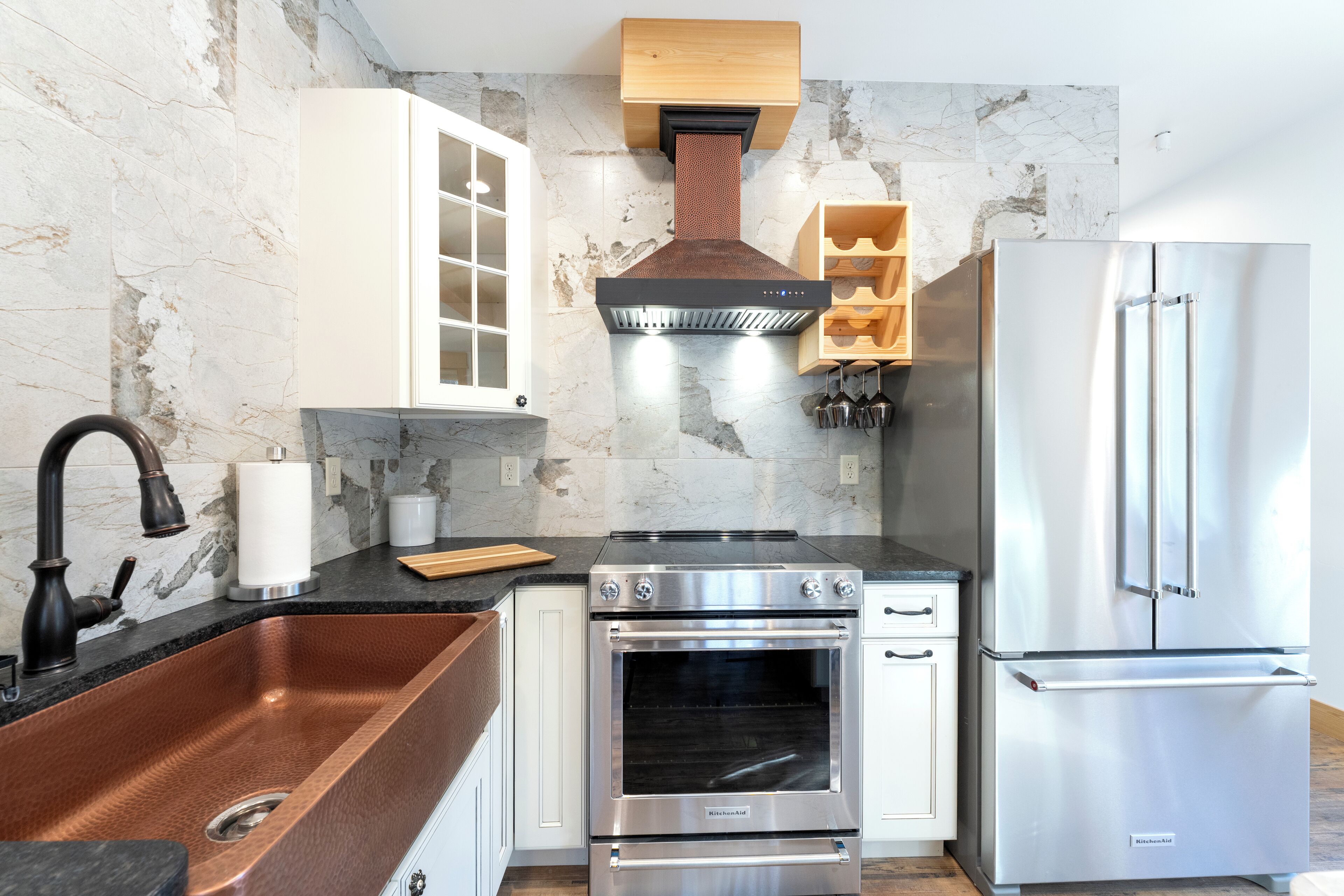 Una cocina moderna con un lavamanos de cobre, electrodomésticos de acero inoxidable, armarios blancos y un salpicadero de piedra.