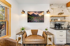 Un salon chaleureux avec un canapé en cuir marron, une table basse en bois et un tableau représentant un paysage de montagne. On y trouve deux tables de chevet avec des lampes, une commode blanche avec des tiroirs et, par la fenêtre, une vue sur la montagne.