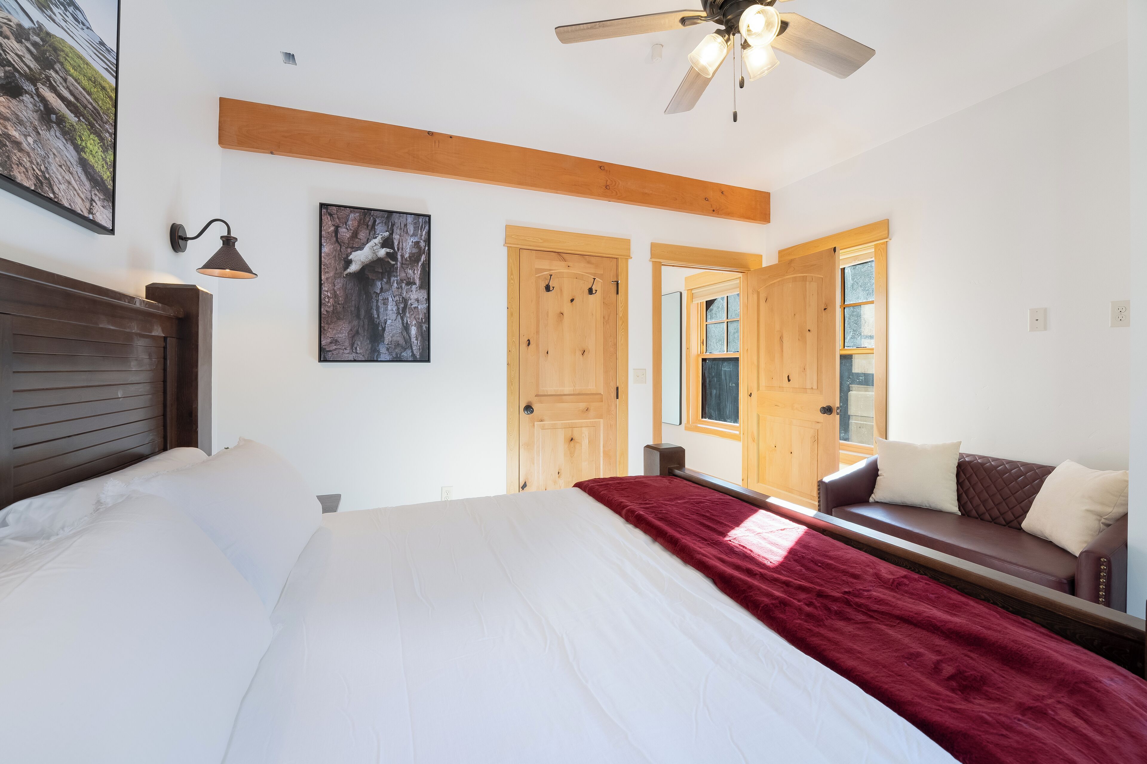A bedroom with a bed, a red throw blanket, a wooden headboard, a ceiling fan, and a wooden bench with cushions.