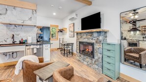 A modern kitchen with a stone fireplace, a dining area with leather chairs, and a large mirror reflecting the kitchen. - New! Lake McDonald Townhome. In Glacier National Park! (West Glacier)
