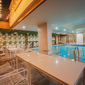 Un área de piscina interior con sillas y mesas de metal blanco, rodeada de paredes con un motivo de hojas verdes.