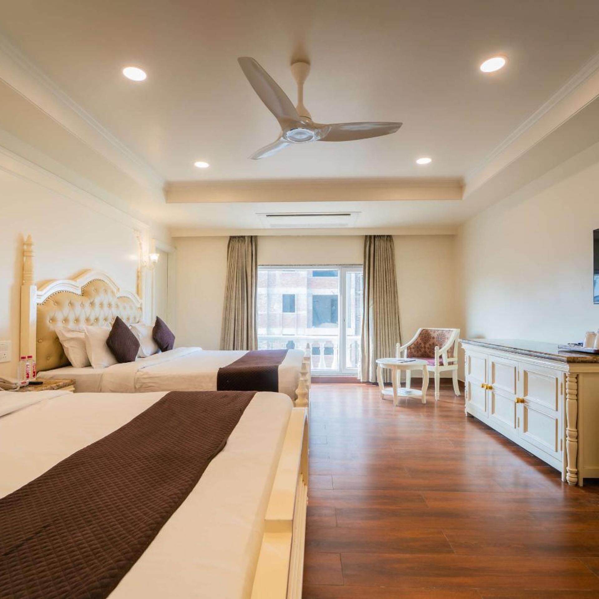 A hotel room with two beds, a ceiling fan, a large window with curtains, a white sofa, and a wooden cabinet.