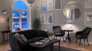 A modern living room with a dark green sofa, a wooden coffee table, and two armchairs. There are framed pictures on the wall, a large mirror, and a ceiling fan. - Hotel Sirius (Kristianstad)