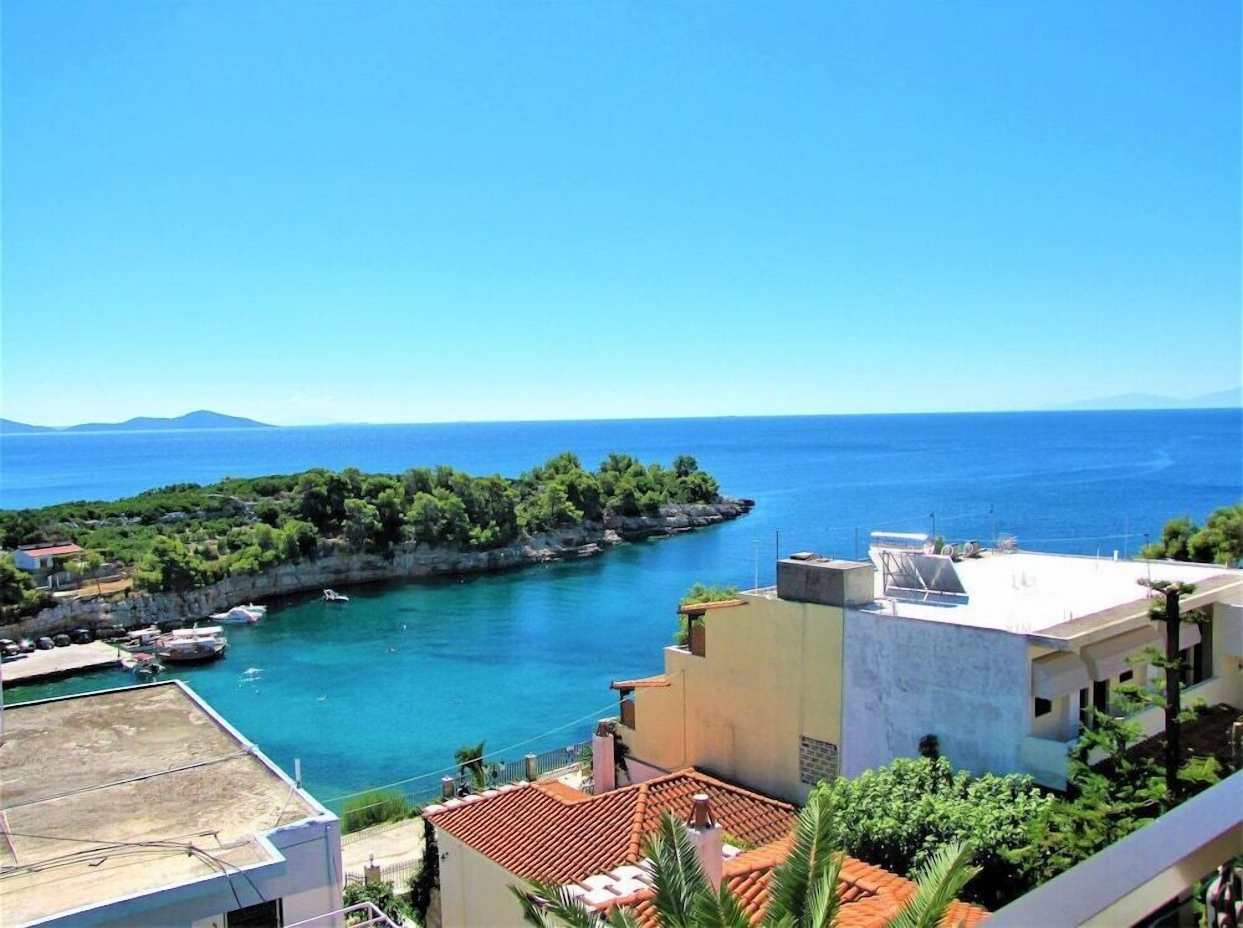 A coastal view with a clear blue sea, a rocky island with trees, and buildings with red-tiled roofs.