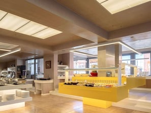 A modern cafe interior with a central yellow counter displaying food items, surrounded by white shelves and a glass display case. - High Beach White - Adults Only (Hersonissos)