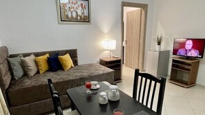 A living room with a sofa, a dining table with chairs, a TV, and a painting on the wall. - Relax Apartments Saranda (Sarandë)