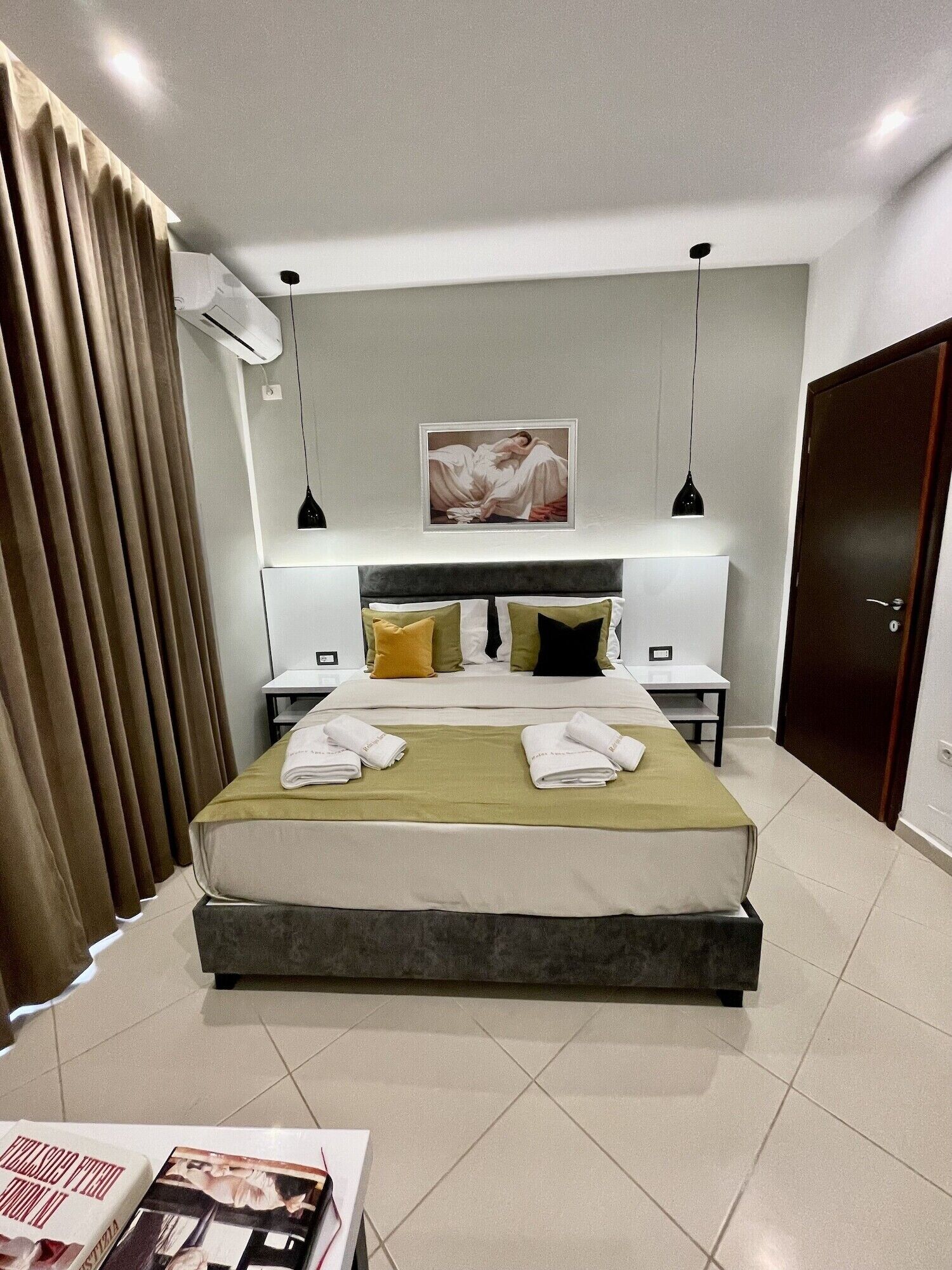 Une chambre d’hôtel moderne avec un grand lit, une table de chevet, une commode, un climatiseur fixé au mur, un luminaire au plafond, un tableau encadré et une porte.